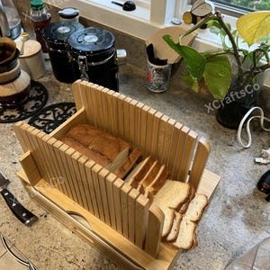May include: A bamboo bread slicer with a loaf of sliced bread. The slicer has vertical slots for guiding the knife and a base to catch the bread. The bread is light brown, and the slicer is made of natural wood.