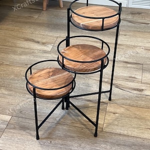 May include: A three-tiered plant stand with a black metal frame and round, wooden shelves. The shelves are made of light brown wood and are surrounded by a black metal railing. The stand has a modern, minimalist design and is suitable for displaying plants or decorative items.