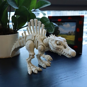 Puede incluir: Figura de esqueleto de dinosaurio color crema con articulaciones y una espalda puntiaguda. El juguete está posado sobre una superficie azul oscuro, con una planta en maceta y una imagen enmarcada en el fondo.