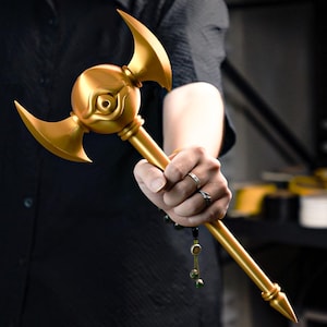 Pode incluir: Um bastão dourado com um orbe central com um design de olho, ladeado por duas lâminas curvas. O bastão é segurado em uma mão, mostrando seus detalhes intrincados e brilho metálico. O design do bastão sugere um tema de fantasia ou cosplay.
