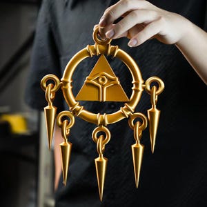 May include: A gold-colored pendant with a circular frame, a central triangle with an eye symbol, and dangling, pointed ornaments. The pendant is held up against a dark background.