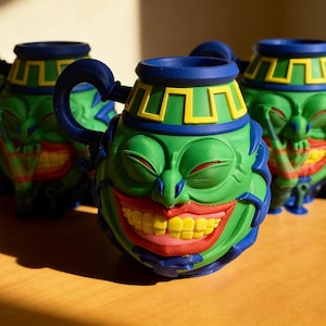 Puede incluir: Tres tazas de cerámica con un diseño de cara de dibujos animados. Las tazas son principalmente verdes con detalles azules y amarillos. La cara tiene una amplia sonrisa con dientes y ojos cerrados.