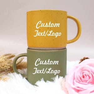 May include: Two stacked ceramic mugs. The top mug is mustard yellow with a curved handle and the text "Custom Text/Logo" in white. The bottom mug is olive green with the same text. The mugs are on a white fluffy surface.