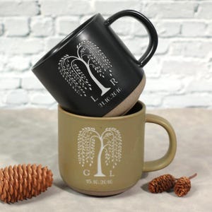 May include: Two ceramic mugs, one black and one tan, stacked. Each mug features a white weeping willow tree design with initials and a date. The black mug has the initials "L R" and the date "21.10.2016". The tan mug has the initials "G L" and the date "15.10.2016".