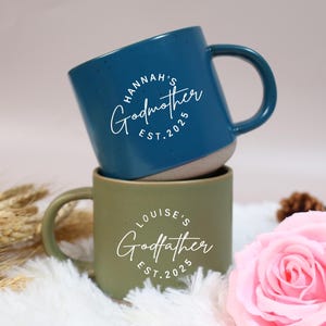 Può includere: Due tazze in ceramica impilate. La tazza superiore è verde acqua con il testo "Hannah's Godmother EST.2025" in corsivo bianco. La tazza inferiore è verde oliva con il testo "Louise's Godfather EST.2025".