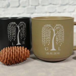 May include: Two ceramic mugs, one black and one tan, each with a white weeping willow tree design. The black mug has the letters "L R" below the tree. The tan mug has the letters "G L" and the date "15.10.2016".