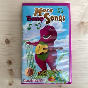 May include: A VHS tape titled "More Barney Songs" featuring Barney the dinosaur playing a guitar. The cover has a blue sky background with musical notes and the text "Never Seen On TV". The tape is encased in a purple plastic case.