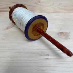 May include: A wooden kite spool with a dark brown handle and a spool of white string. The spool has a dark brown rim, a gold band, and a blue outer ring. The spool is lying on a light wood surface.