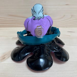 May include: A figurine of a character with a light blue head and purple torso, seated on a black base with teal accents. The figurine has a clear plastic piece attached to the front.