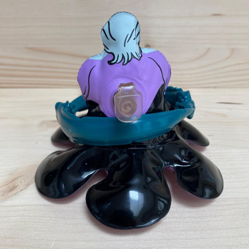 May include: A figurine of a character with a light blue head and purple torso, seated on a black base with teal accents. The figurine has a clear plastic piece attached to the front.