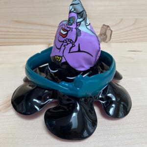 May include: A black and teal Ursula-themed toy with a purple head and a teal band around the base. The toy has a cartoon image of Ursula on the head. The base has four black, glossy, petal-shaped elements.