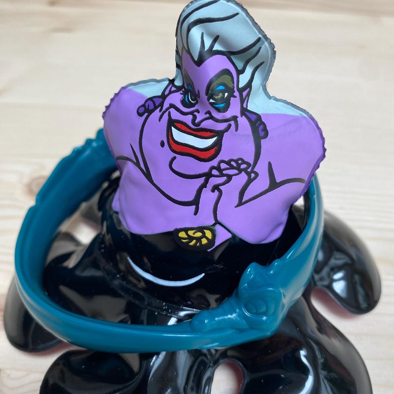 May include: A figurine of Ursula, the sea witch from Disney's The Little Mermaid. The figure has a purple face with a sinister grin, white hair, and a black dress. A teal-coloured collar surrounds the figure.
