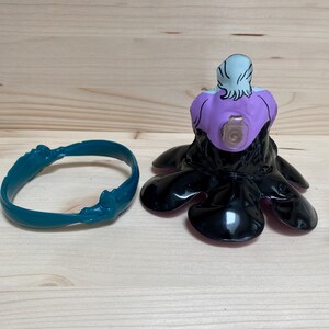 May include: A teal bracelet and a black and purple Ursula figurine. The figurine has a purple face and white hair. The figurine is in the shape of an octopus. The bracelet is a solid teal colour.