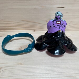 May include: A purple and black Ursula figurine from The Little Mermaid, with a teal bracelet. The figurine features Ursula's face and tentacles. The bracelet is teal and has a wave-like design.