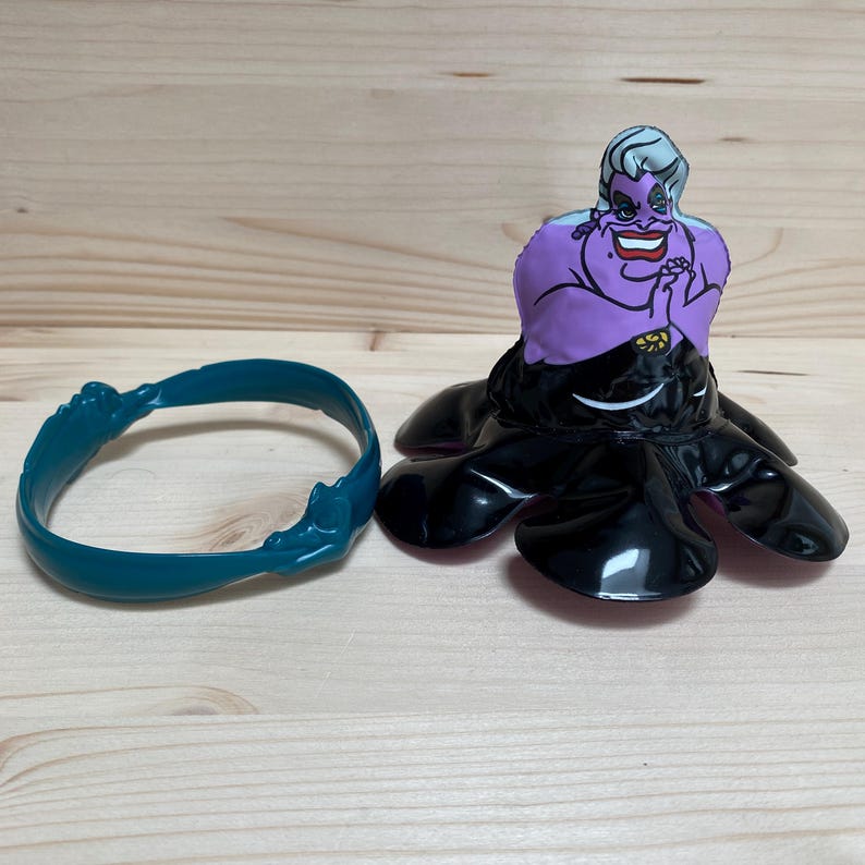 May include: A purple and black Ursula figurine from The Little Mermaid, with a teal bracelet. The figurine features Ursula's face and tentacles. The bracelet is teal and has a wave-like design.