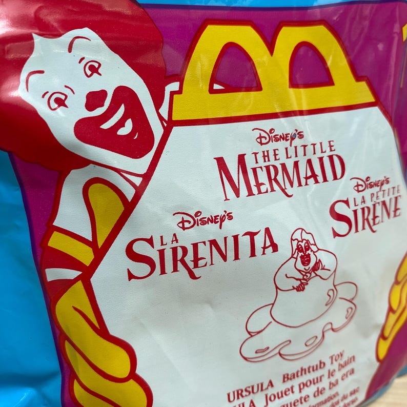 May include: A colourful McDonald's Happy Meal bag featuring Ronald McDonald and Disney's The Little Mermaid. The bag has text in English, French, and Spanish, and includes an image of Ursula.
