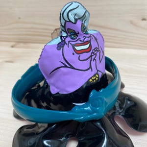 May include: A purple Ursula the Sea Witch figure from Disney's The Little Mermaid. The figure has a teal collar and a black base. The character has white hair, red lipstick, and a sinister smile. The figure is a collectible item.