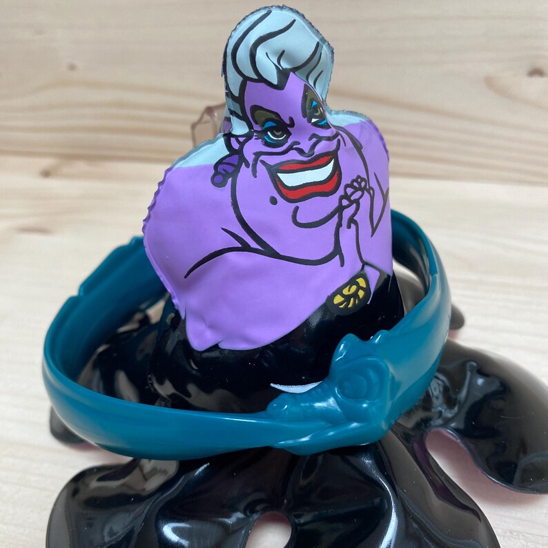 May include: A purple Ursula the Sea Witch figure from Disney's The Little Mermaid. The figure has a teal collar and a black base. The character has white hair, red lipstick, and a sinister smile. The figure is a collectible item.