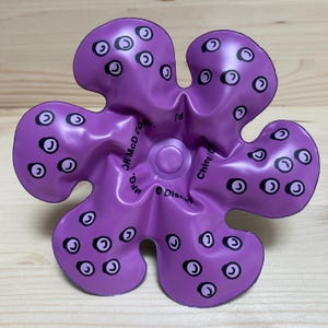 May include: A purple, flower-shaped toy with black and white circular designs. The toy has a central circular feature and text that reads "&copy; Disney" and "China/C".