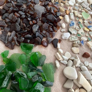 May include: An assortment of natural beach finds, including sea glass in shades of brown and green, along with small pebbles and fragments of pottery. The items are arranged on a light tan surface, showcasing their varied textures and colors.