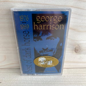 May include: A cassette tape of George Harrison's "Best of Dark Horse" album, spanning 1976-1989. The cover features a blue background with Harrison's image and gold text. Includes a sticker with additional information.