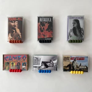 May include: Six cassette tapes displayed on a white wall. The tapes feature album art and text, including "YOUNG M.C.", "METALLICA", and "DINOSAUR JR". Each tape is held by a row of colorful, rounded holders.