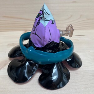 May include: A black, flower-shaped toy with a teal ring and a purple and white fabric cone. The cone features a cartoon character design. A clear plastic piece extends from the side of the cone.