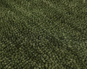 Pode incluir: Close-up de um tapete verde-oliva texturizado. O tapete tem uma pilha densa e em laço, criando uma aparência macia e felpuda. A cor é um verde suave e terroso, com variações sutis de tom.