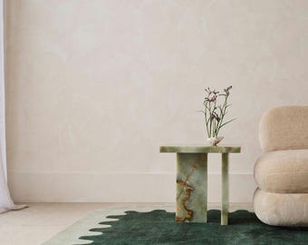 May include: A modern living room scene featuring a green and light green wavy-edged rug. A small green marble side table holds a vase of flowers. A beige armchair is partially visible in the background.