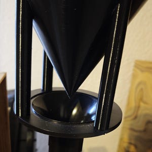 Könnte beinhalten: Ein schwarzes, geometrisches Objekt mit konischer Form und kreisförmiger Basis. Das Objekt wird von vier vertikalen Stäben getragen und sitzt auf einer zylindrischen Struktur. Das Design ist modern und industriell, mit klaren Linien und minimalistischer Ästhetik.