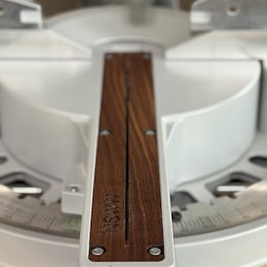 May include: Close-up of a miter saw with a wooden insert. The insert has the text "AMSN" engraved on it. The saw is primarily white with a silver blade guard and a measurement scale.