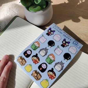 May include: A sheet of clear vinyl stickers featuring various cartoon characters in a variety of colors. The stickers are on a light blue background and are placed on an open notebook. A small potted plant is in the background.
