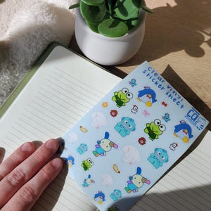 May include: A sheet of clear vinyl stickers with cartoon characters, including a frog, penguin, and bunny, on a light blue background. The stickers are on a notebook with lined pages. A small potted plant is in the background.