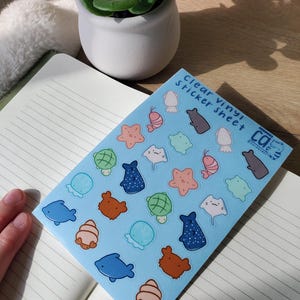 May include: A clear vinyl sticker sheet featuring various sea creatures in a light blue background. The stickers include a blue dolphin, brown bear, pink starfish, and green turtle. The sheet also has the text "Clear Vinyl Sticker Sheet".