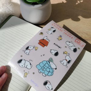 May include: A clear vinyl sticker sheet featuring Snoopy in various poses and outfits. The sheet includes Snoopy in a blue coat, a red doghouse, and other designs. The text "Clear Vinyl Sticker Sheet" and "esnupi" are visible.