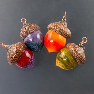 May include: Four acorn-shaped pendants featuring smooth, glossy, multi-colored glass bodies in red, purple, orange, and yellow. Each pendant is topped with a textured, brown, realistic acorn cap and includes a small copper wire loop for hanging.