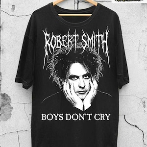 May include: Black t-shirt featuring a graphic of Robert Smith with the text "ROBERT SMITH" above his image and "BOYS DON'T CRY" below. The shirt is hanging on a wooden hanger against a textured wall.