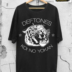 Könnte beinhalten: Schwarzes T-Shirt mit einem weißen Tiger-Motiv und dem Text "Deftones Koi No Yokan". Das T-Shirt hängt an einem Holzbügel vor einer strukturierten Wand. Das T-Shirt hat auch das Logo "EVC apparel".