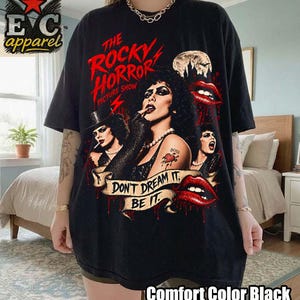 Op de afbeelding: Een zwart t-shirt met een grafische print van The Rocky Horror Picture Show. Het ontwerp bevat personages, rode lippen, een hart-tattoo en de tekst: The Rocky Horror Picture Show, Don't Dream It, Be It. Het shirt heeft een ronde hals.
