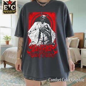 May include: Dark gray Comfort Color Graphite t-shirt featuring a red and black graphic of a horned figure in a white robe. The graphic is set against a red background with the word "SINISTER" in red. The shirt has short sleeves.