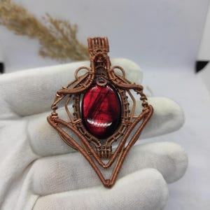 May include: A copper pendant with a striking, oval red gemstone. The gemstone is encased in an elaborate wire-wrapped design, with geometric patterns and fine details. The pendant has a loop at the top for a chain. The pendant is approximately 4 cm in length.