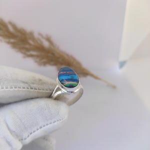 May include: A silver ring with an oval gemstone displaying a colourful pattern of blues, reds, and greens. The ring has a simple, polished silver band. The gemstone's colours provide a striking contrast against the silver.