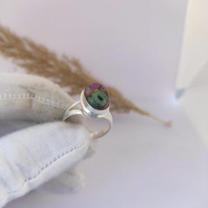 May include: A silver ring featuring an oval-shaped gemstone with a mix of colors, including green, pink, and brown. The ring has a split shank design. The gemstone is the focal point, set in a polished silver band.