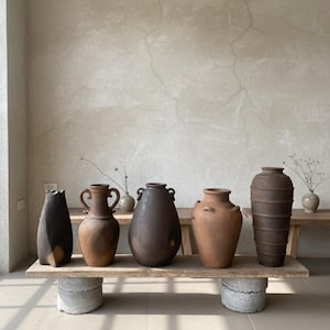 May include: A display of several ceramic vases in different shapes and sizes, arranged on a weathered wooden shelf. The vases are in shades of brown and terracotta, some with handles. The shelf is supported by concrete blocks.