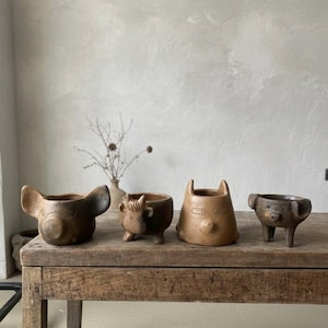 May include: Four brown ceramic animal-shaped planters. The planters are shaped like a dog, a bull, a cat, and a pig. They are arranged on a rustic wooden table. The background is a neutral-toned wall with a vase of dried flowers.