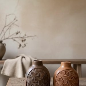 May include: Two ceramic vases with a textured, wave-like pattern. One vase is a dark brown, the other is a lighter terracotta color. The vases are on a wooden table, with a neutral background.