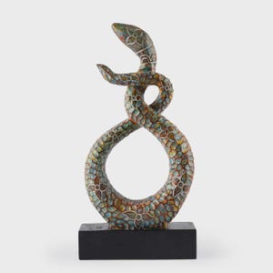 Puede incluir: Escultura decorativa de serpiente con un diseño entrelazado único. La escultura presenta un patrón detallado de motivos florales en tonos verdes, azules y dorados. Está montada sobre una base rectangular negra.