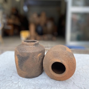May include: Two small, handmade terracotta pots with a rustic, textured finish. One is upright with a narrow neck, the other is on its side with a circular opening. The pots are a warm brown color with darker patches.