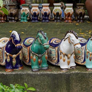 May include: A collection of ceramic horse ornaments in shades of red, blue, green, white, and beige. Each horse features a detailed saddle and decorative elements. The ornaments are displayed outside.