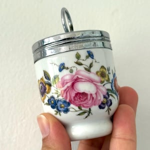 May include: White porcelain egg cup with a floral design featuring a large pink rose and smaller blue, yellow, and purple flowers. The cup has a silver-colored metal rim and handle.